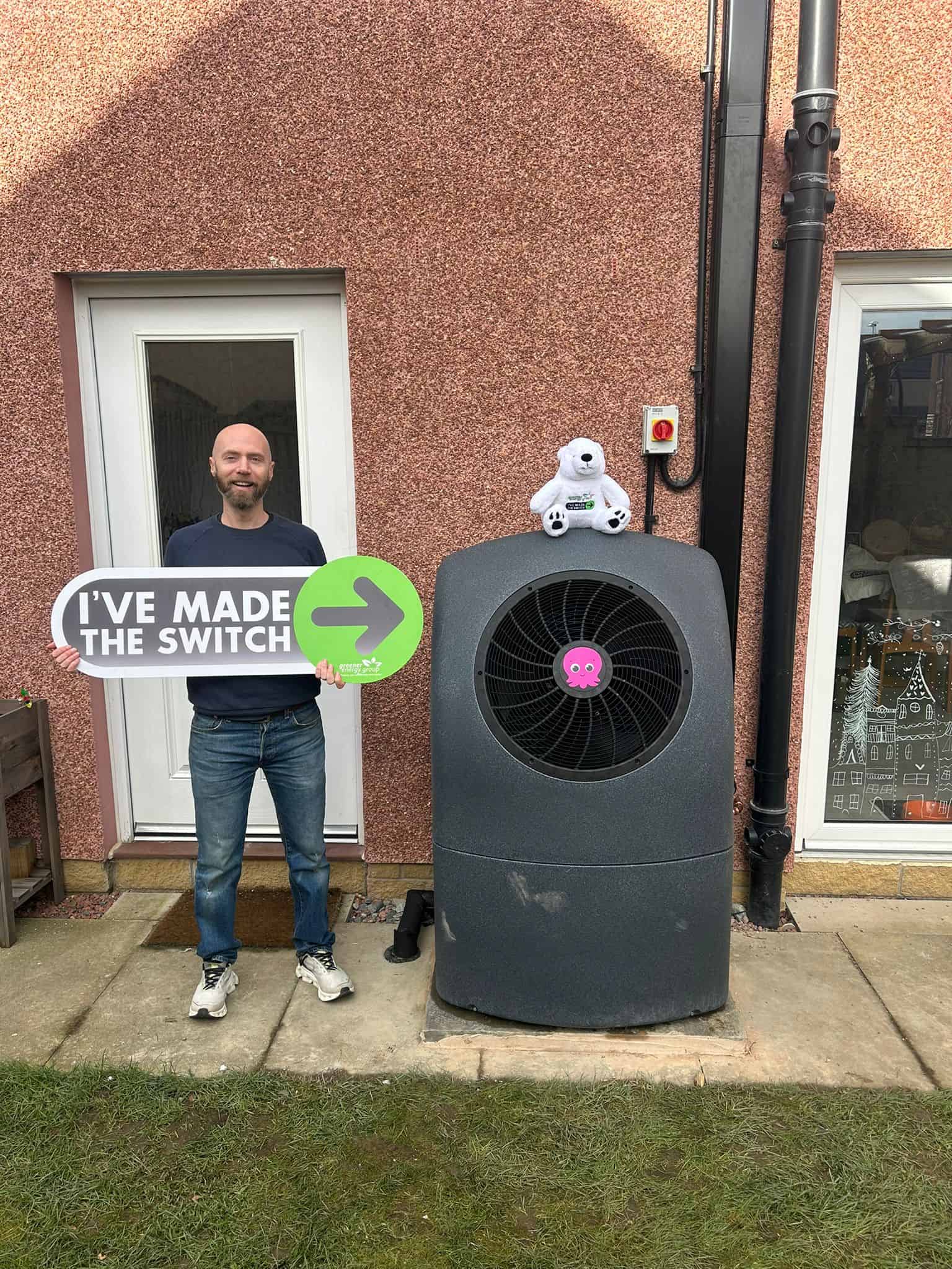 Octopus Cosy Heat Pumps 9 Man standing next to a heat pump with a "Made the Switch" sign outside a house.